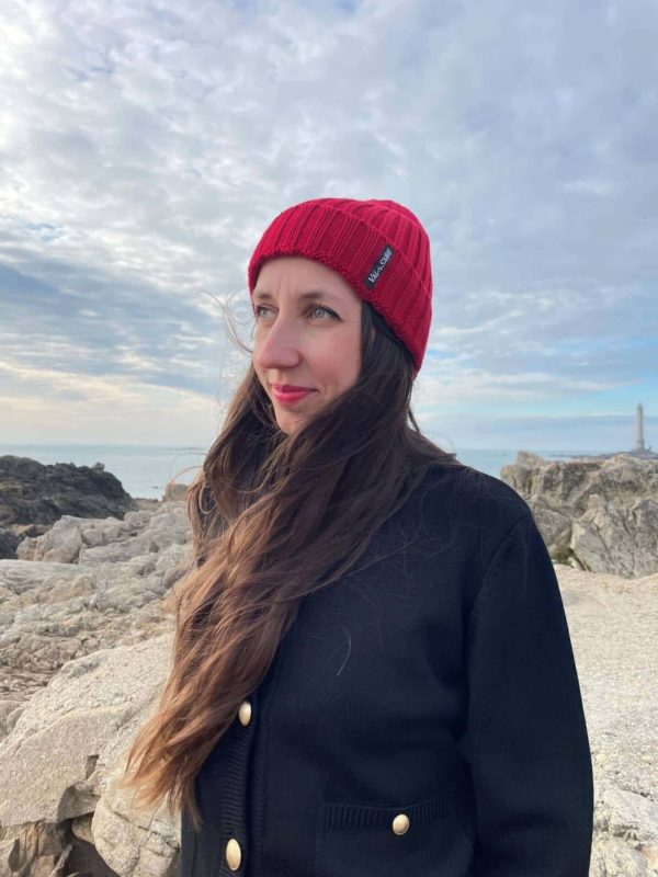 Bonnet Brick couleur rouge porté par une femme qui regarde vers la gauche avec en fond les rochers et la mer