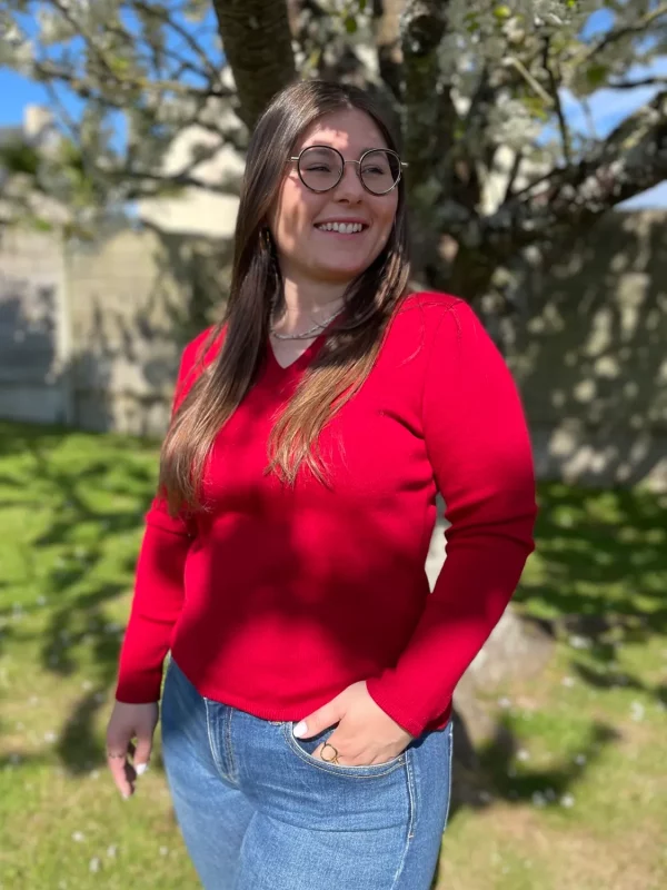 Pull col V en 100% laine mérinos extrafin rouge sombre porté avec un jean par une femme dans un jardin ensoleillé