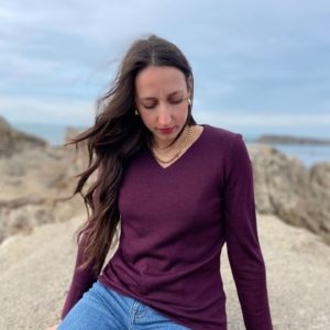Pull col V couleur aubergine porté avec un jean par une femme assise sur un rocher.