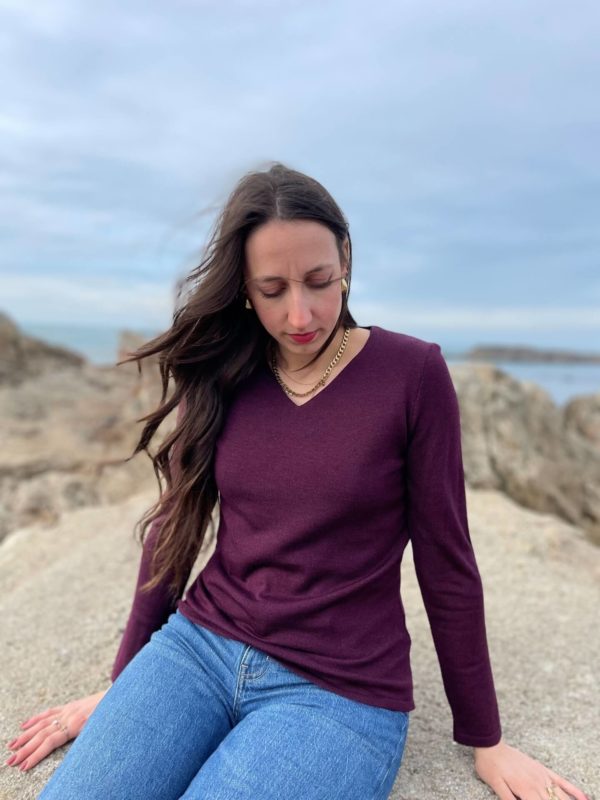 Pull col V couleur aubergine porté avec un jean par une femme assise sur un rocher.