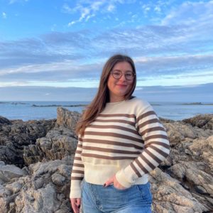 Pull en 100% mérinos extrafin écru avec des rayures couleur châtaigne porté avec un jean par une femme debout sur des rochers avec la mer en fond