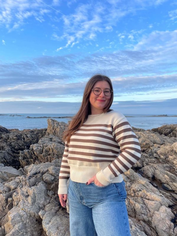Pull en 100% mérinos extrafin écru avec des rayures couleur châtaigne porté avec un jean par une femme debout sur des rochers avec la mer en fond